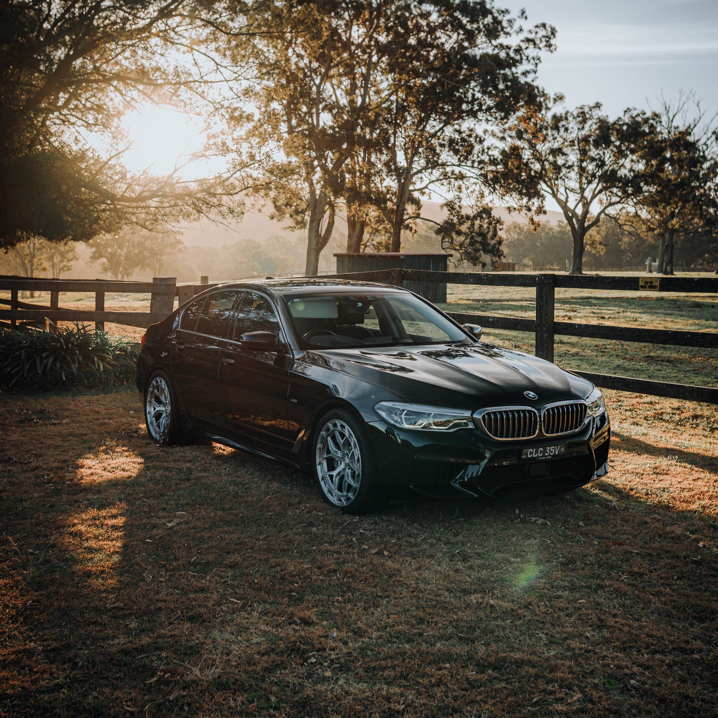 BMW car parked on a grassy field with trees and a wooden fence in the background during sunset.